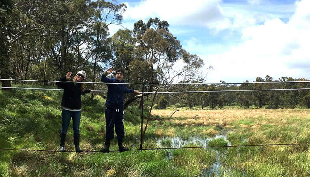 Two people walk across ropes suspended above a waterhole in bushland, looking to camera 